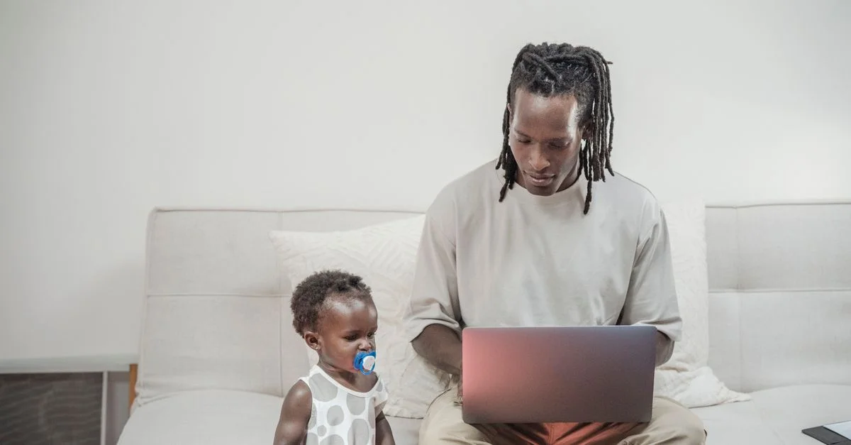 Un père travaille sur un ordinateur portable à la maison pendant que sa fille en bas âge est assise à côté de lui, mettant en avant l'équilibre familial.