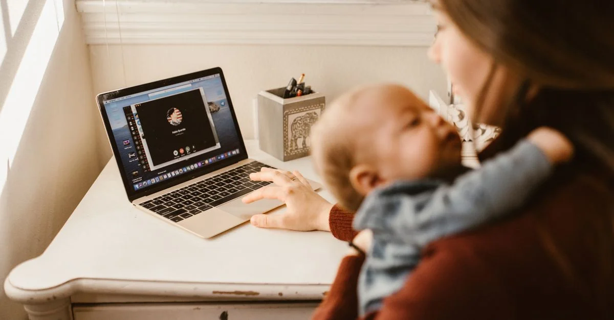 Une mère tenant son bébé tout en faisant un appel vidéo sur un ordinateur portable dans un cadre domestique confortable.