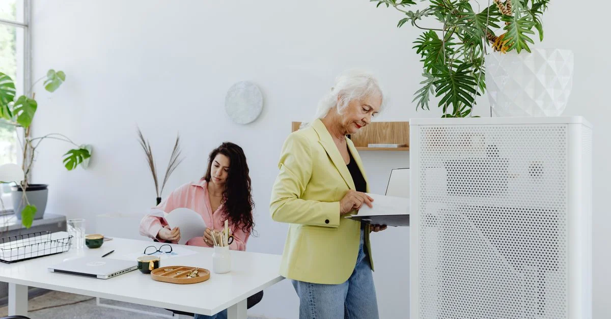 Femme d'affaires senior en blazer vert travaillant avec une collègue dans un bureau moderne.