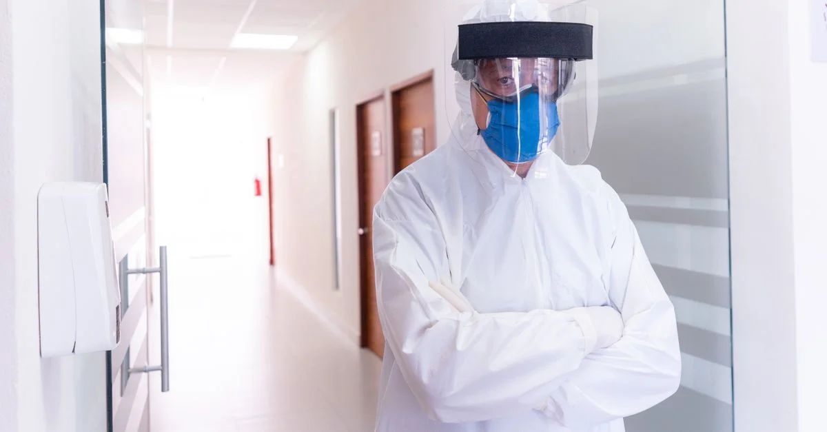 Professionnel de la santé en tenue complète d'EPI se tient dans un couloir d'hôpital, portant un masque facial et une visière.