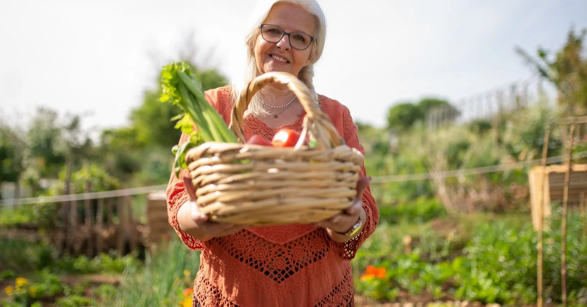 Femme âgée souriante avec un panier de légumes frais dans un jardin extérieur vibrant.